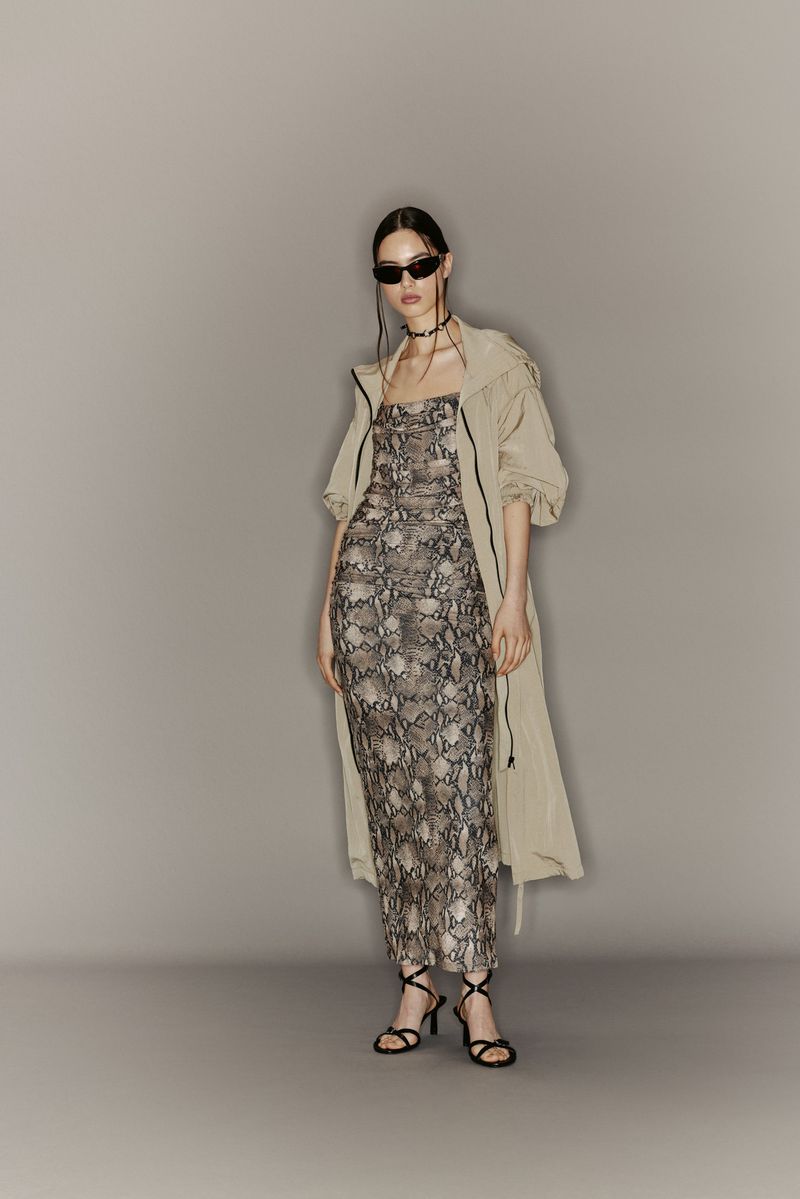 Fashionable woman in snakeskin dress and beige trench coat posing in studio. She wears sunglasses and strappy heels, looking elegant.