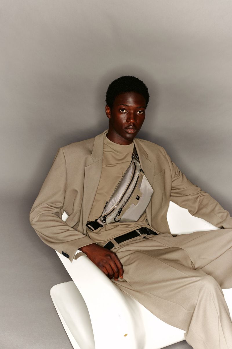 Young adult African-descent male model in a beige suit with a beige fanny pack, seated indoors against a grey background in portrait studio.