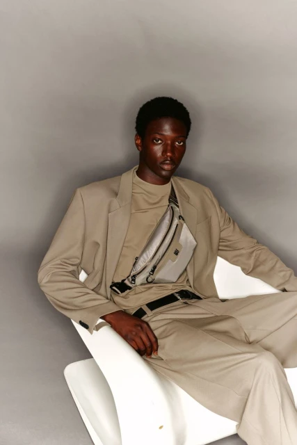 Young adult African-descent male model in a beige suit with a beige fanny pack, seated indoors against a grey background in portrait studio.