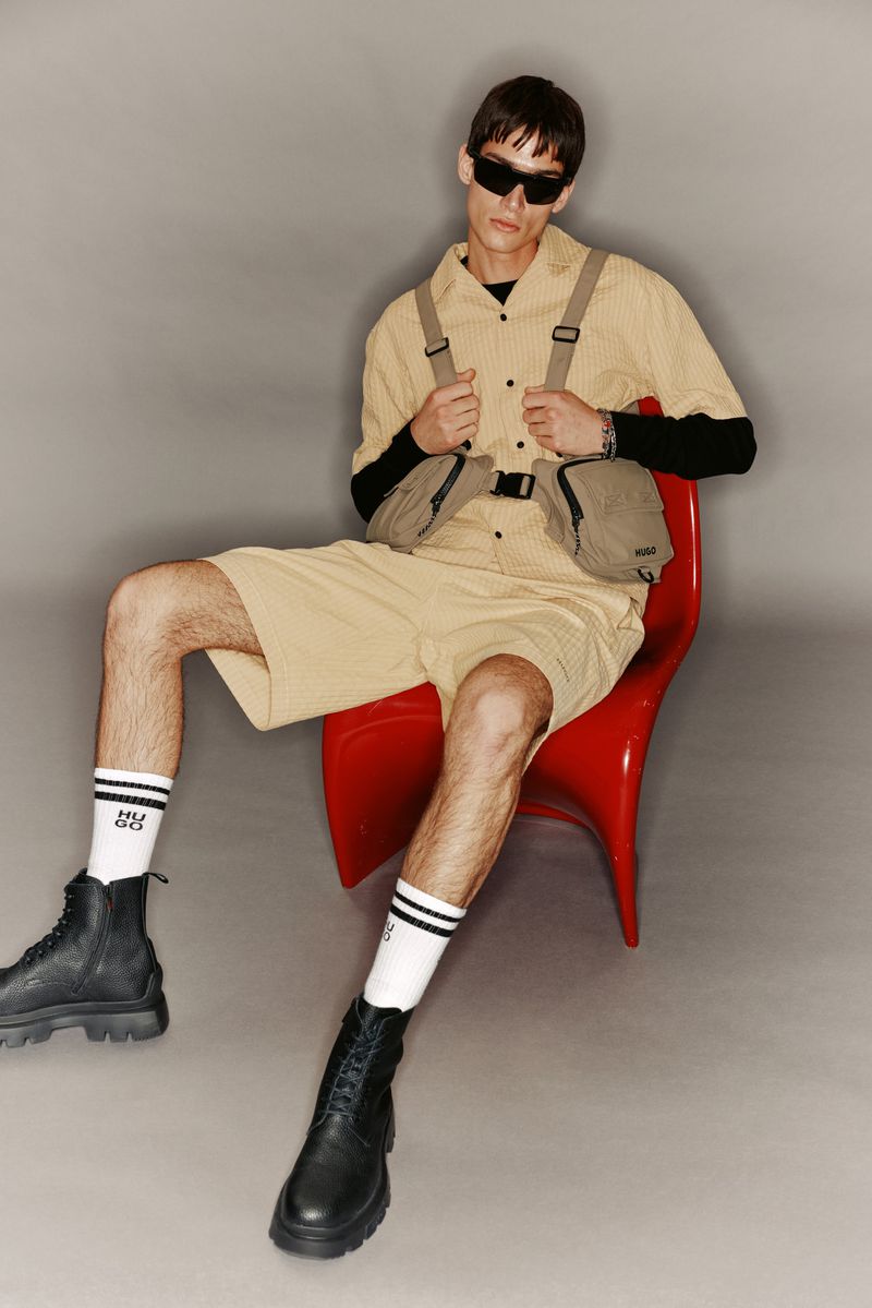 Man seated in a red chair wears a tan collared shirt, shorts, black boots, and sunglasses, posing against a gray background in a studio setting.