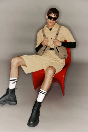 Man seated in a red chair wears a tan collared shirt, shorts, black boots, and sunglasses, posing against a gray background in a studio setting.