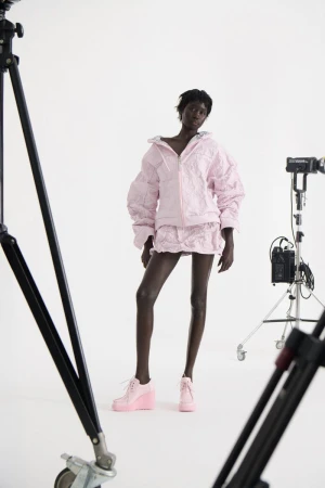 Black model posing in a pastel pink textured jacket, mini skirt, and matching wedge shoes against a white studio background with equipment.