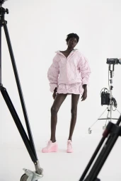 Black model posing in a pastel pink textured jacket, mini skirt, and matching wedge shoes against a white studio background with equipment.