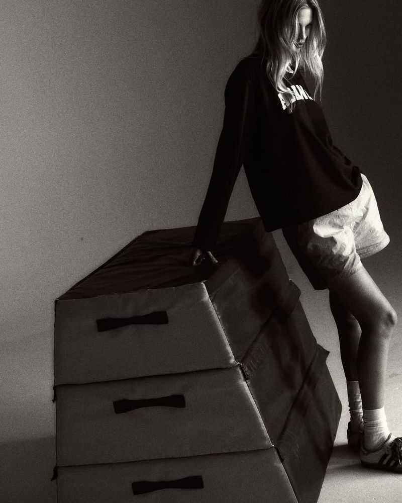 Monochrome shot of a blonde woman in casual athletic wear, leaning on a three-tiered gymnastic vault box in a studio setting.