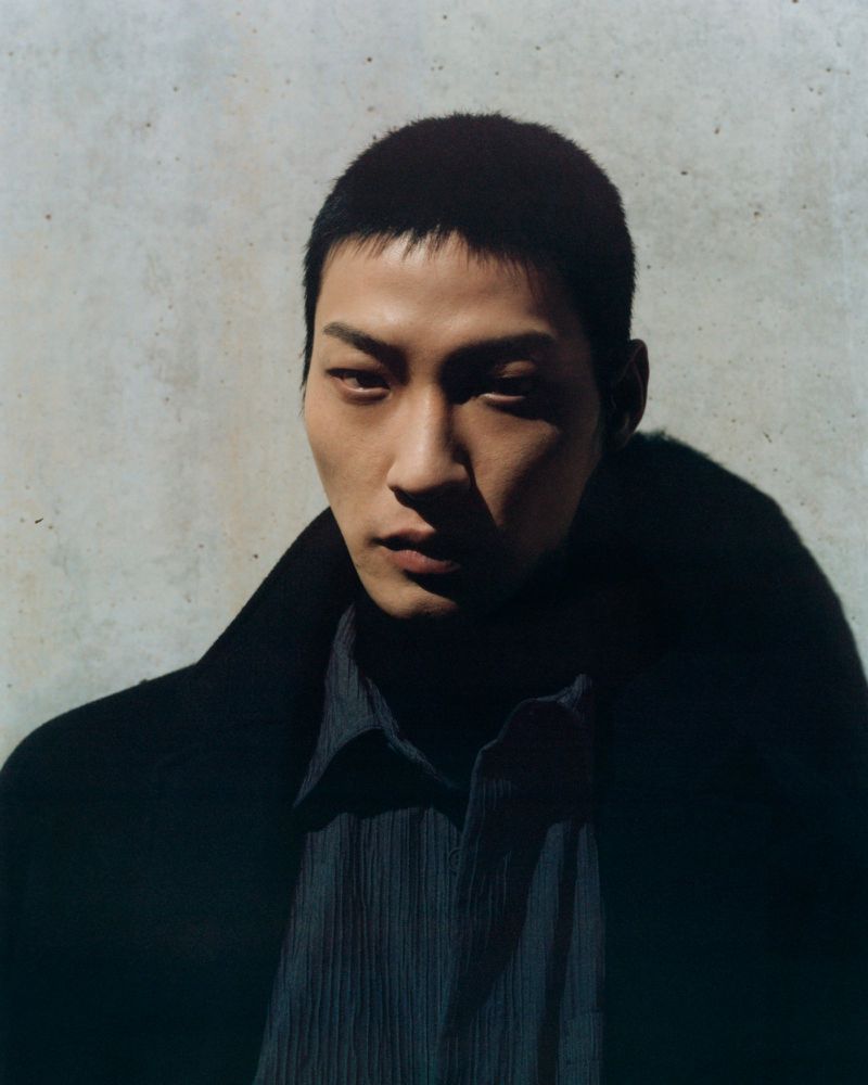 Portrait of an adult Asian male in a dark coat and collared shirt standing in front of a light grey wall in soft light.