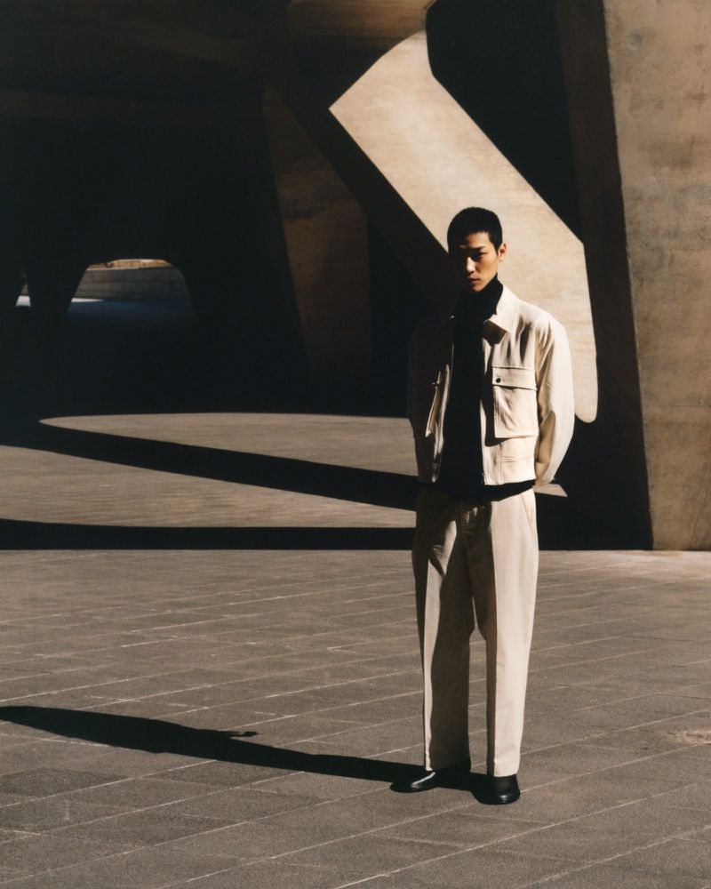Young Asian man in a cream-colored outfit stands against an abstract concrete background in shadows and light, creating a modern portrait.
