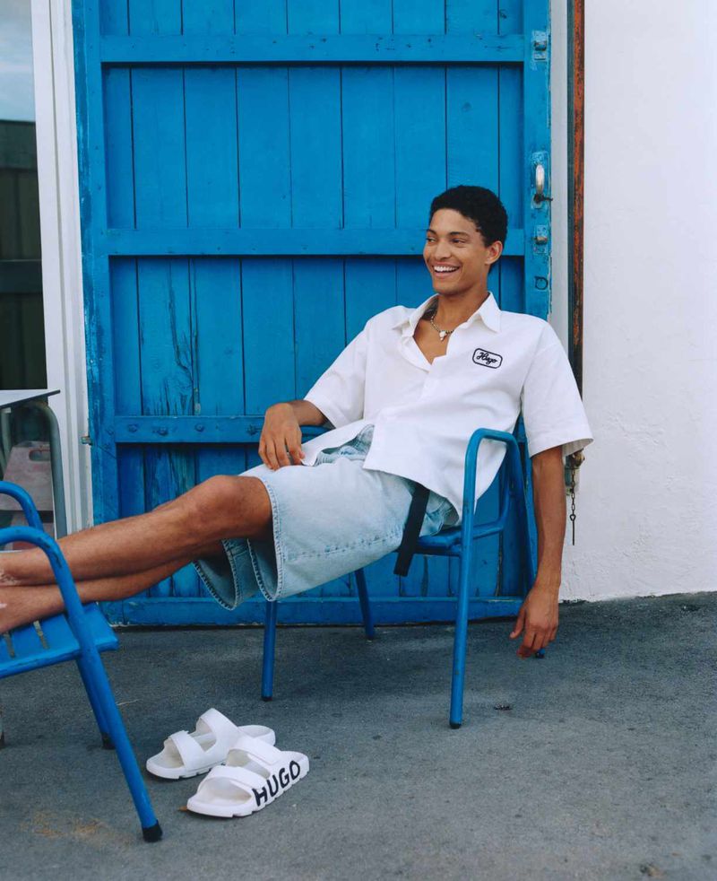Smiling adult man in casual clothes sits on a blue chair outdoors with white sandals with logo, against a blue door and white wall.