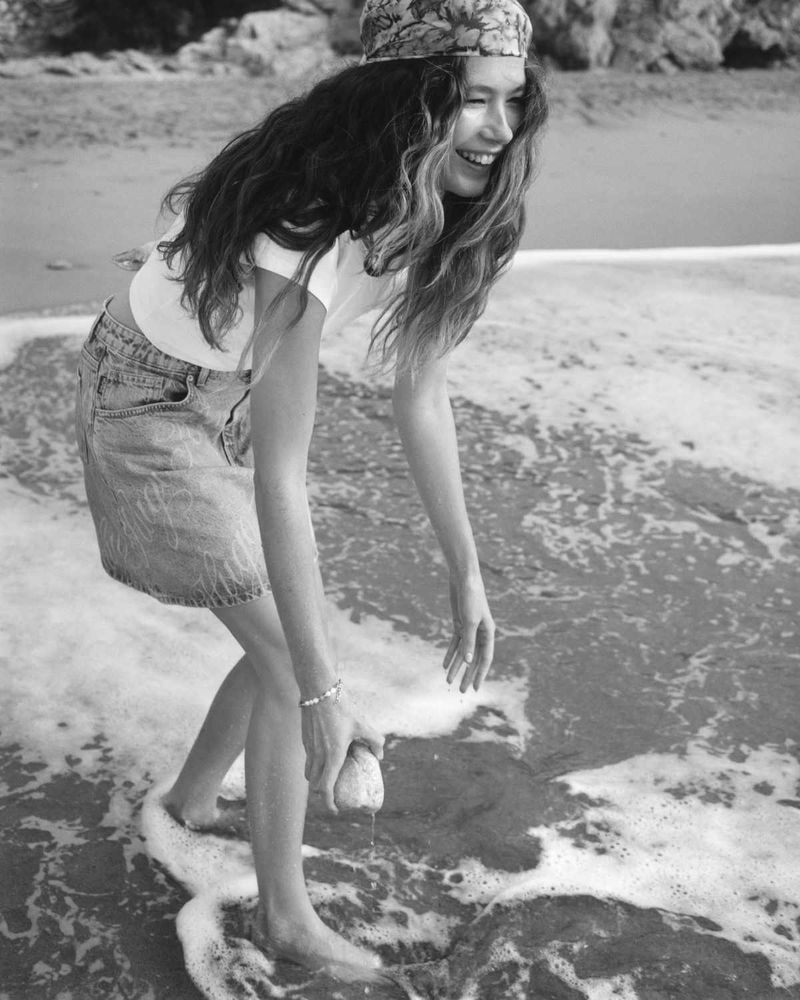 A woman smiles on a beach, wearing a headscarf, t-shirt, and denim skirt while holding a rock in the ocean water, captured in black and white.