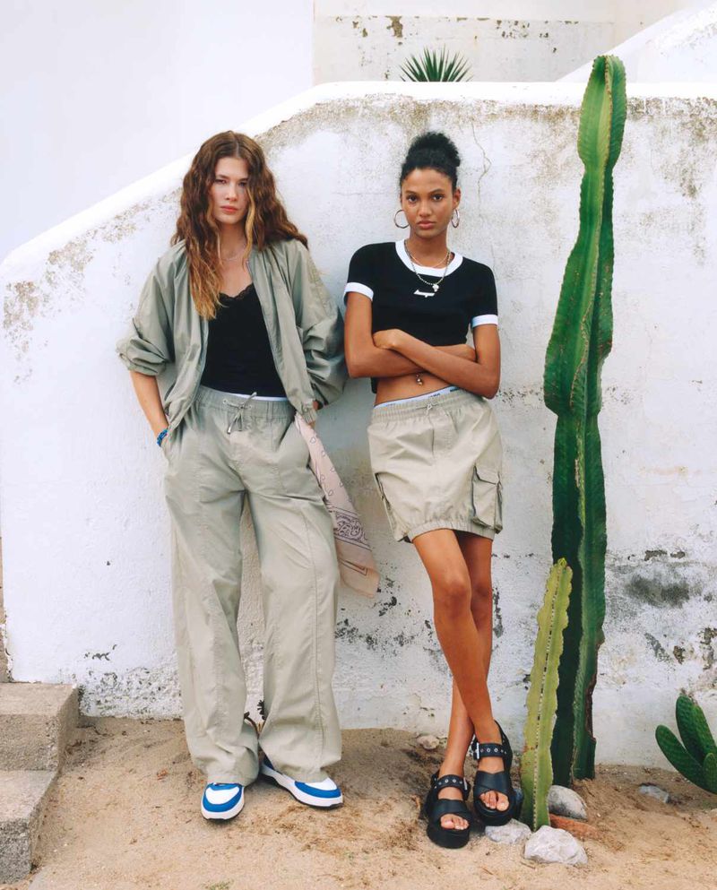Two women pose near a tall cactus against a white wall, wearing casual fashion. One wears loose pants and the other a skirt.