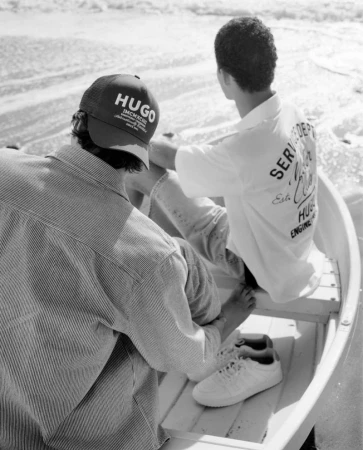 Two men in a small boat on a black and white beach scene. One wears a HUGO cap and the other a white shirt with graphic text.