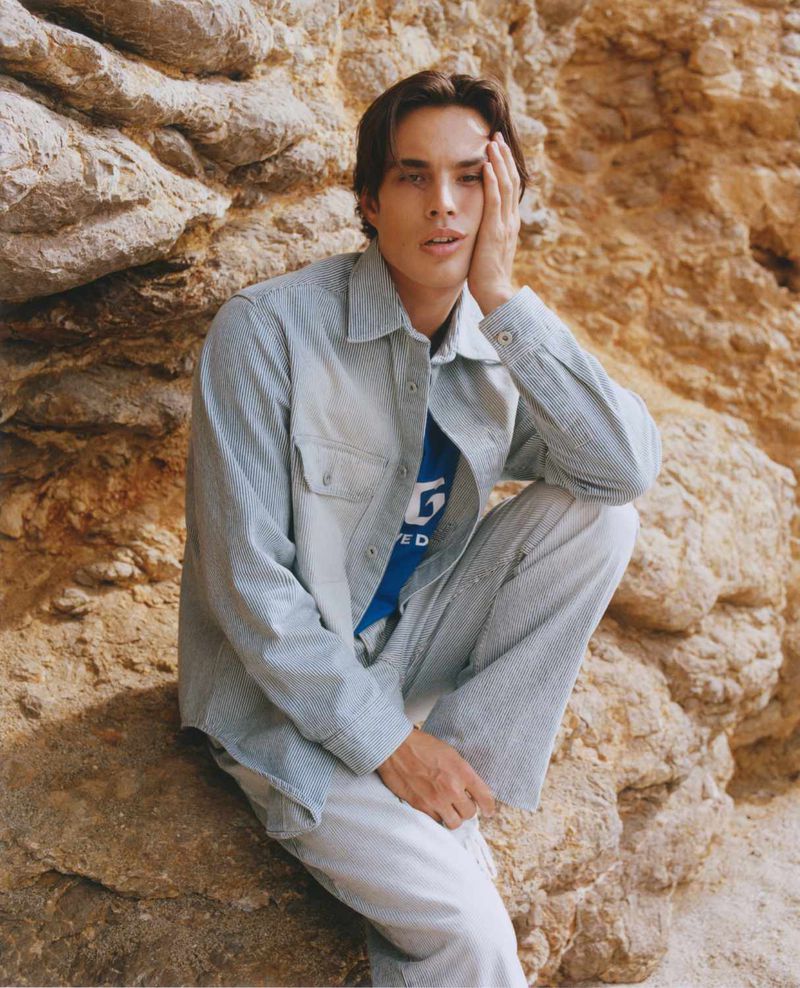 An adult male with dark hair poses with his hand near his face, wearing a striped jacket and pants, seated against a rocky backdrop.