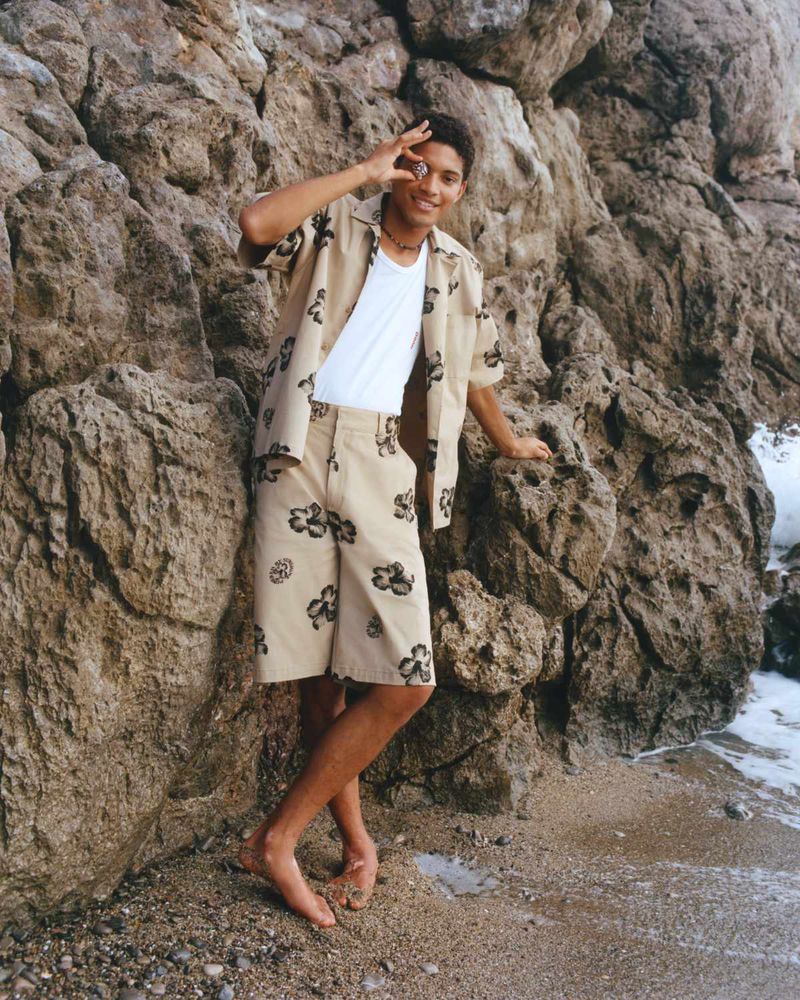 A young adult male stands on a sandy beach in shorts and a patterned shirt, posing with one hand near his eye by a rock formation.