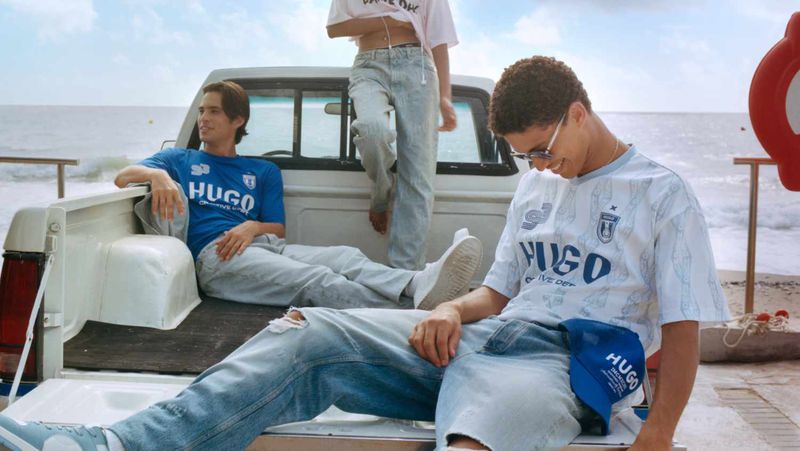 Three young adults pose in the back of a white pickup truck near a beach with ocean views, wearing Hugo branded apparel.