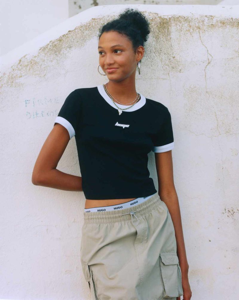 A young adult woman poses outdoors in a black short-sleeved top and khaki skirt, showcasing a casual yet stylish fashion look in natural light.