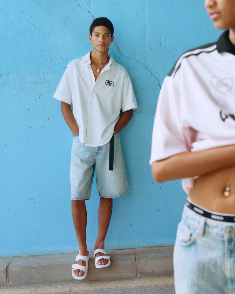 A male model poses in a white shirt and denim shorts against a blue wall, while a second model appears in soft focus on the right.