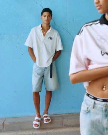 A male model poses in a white shirt and denim shorts against a blue wall, while a second model appears in soft focus on the right.
