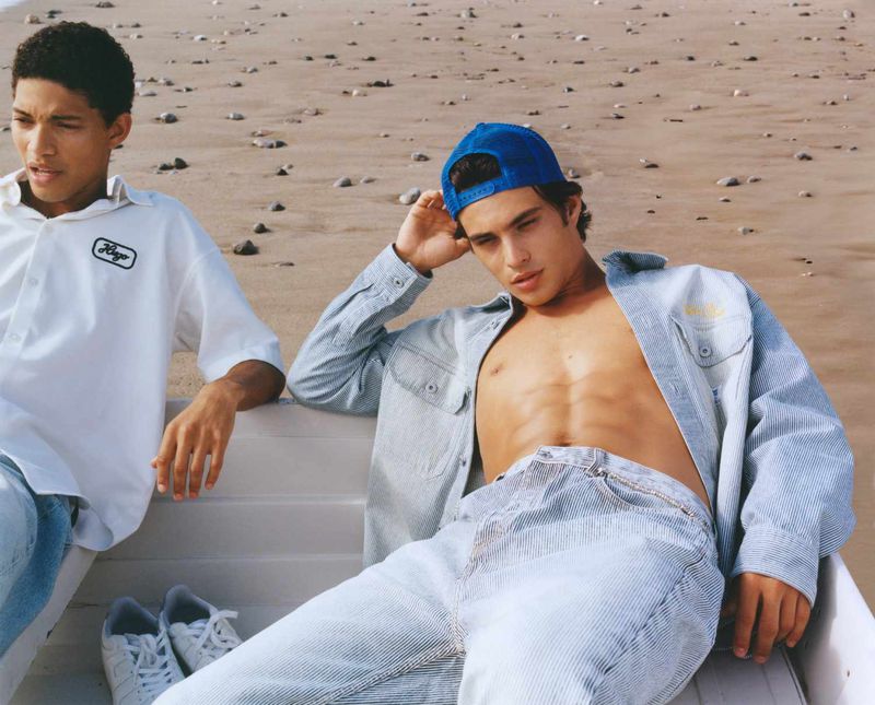 Two young adult men pose on a sandy beach. One wears a blue baseball cap, light blue jeans and jacket, and the other wears white clothing.