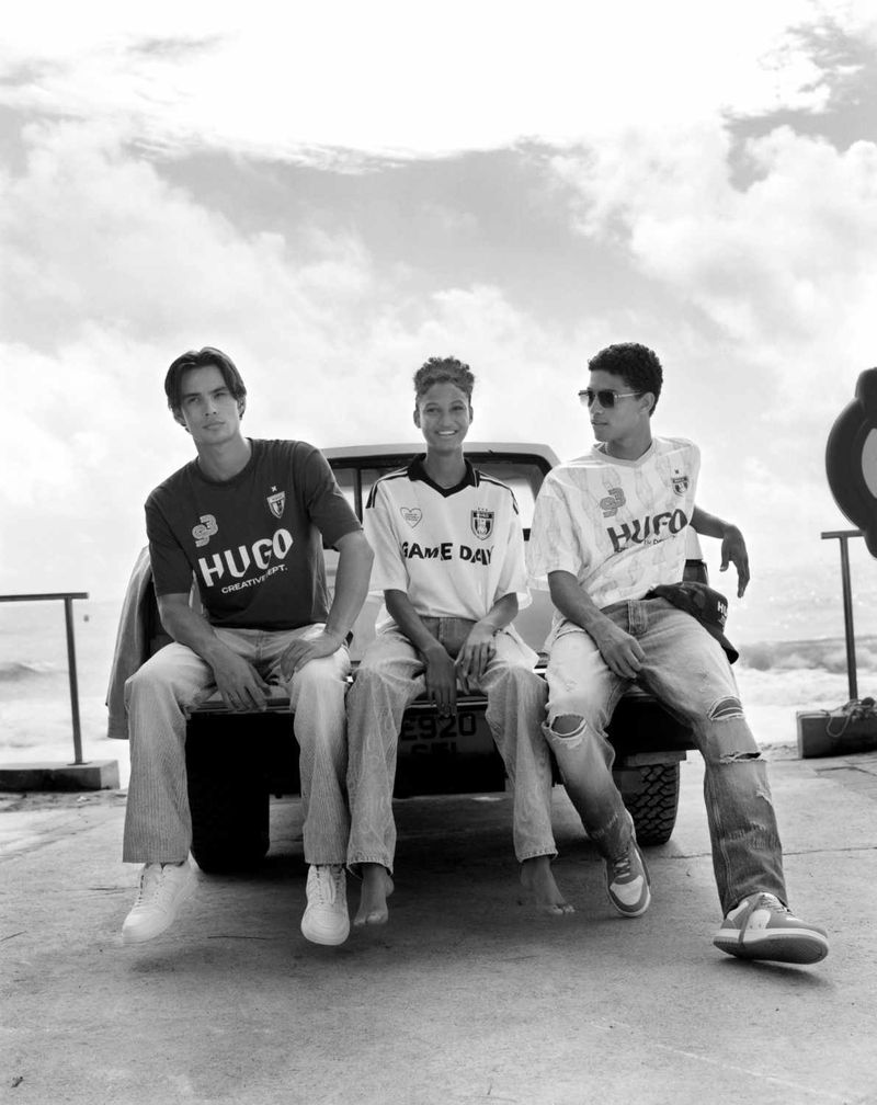 Three young adults in vintage soccer jerseys sit on a truck bed with ocean and cloudy sky in the background in a black and white fashion shot.