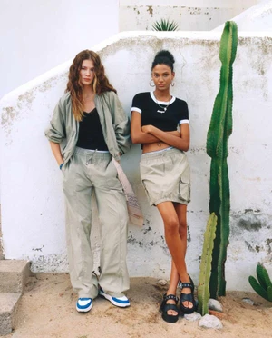 Two models posing against a white rendered wall near a tall cactus for the HUGO SS25 campaign.