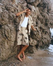 Young man standing on a sandy beach leaning against rocky cliffs wearing patterned beige shirt and shorts