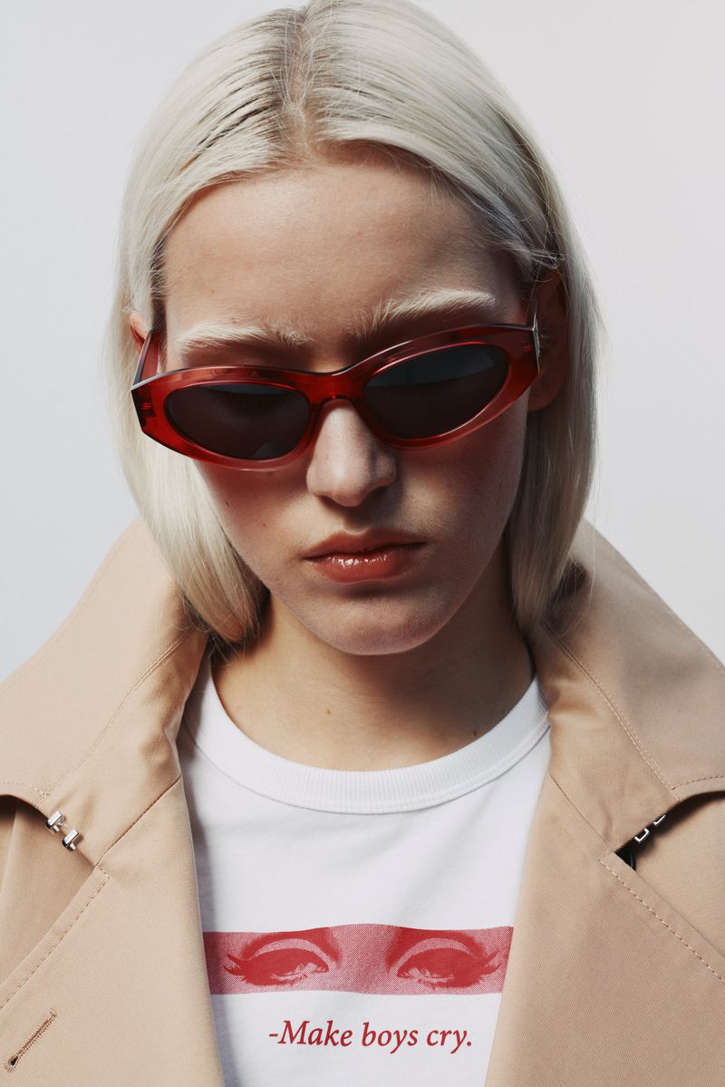 Fashion portrait of a blonde woman wearing red sunglasses and a trench coat in a studio setting with a white background.