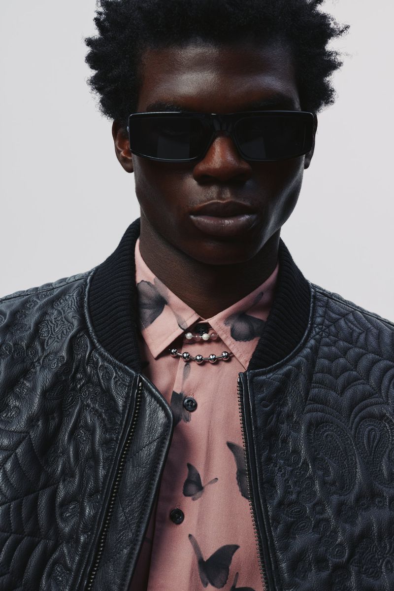 Headshot of a dark-skinned adult male wearing sunglasses and a quilted bomber jacket over a collared shirt with a butterfly pattern.