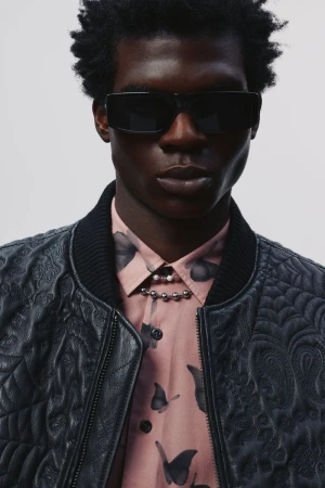 Headshot of a dark-skinned adult male wearing sunglasses and a quilted bomber jacket over a collared shirt with a butterfly pattern.