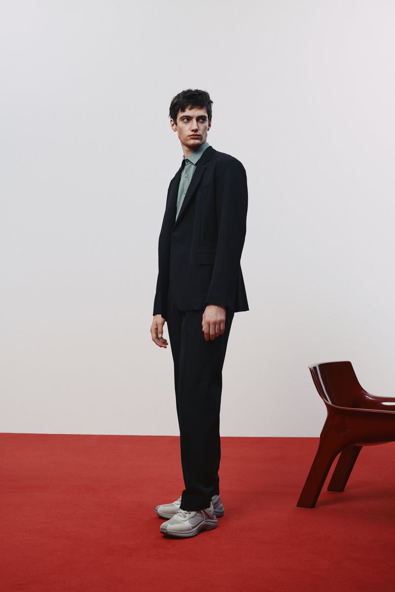 A man in a black suit and sneakers stands on a red floor in front of a white wall, next to a unique red chair in a fashion pose.