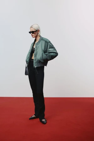 Model wearing a green bomber jacket, black trousers, and sunglasses standing on a red floor in front of a white wall.