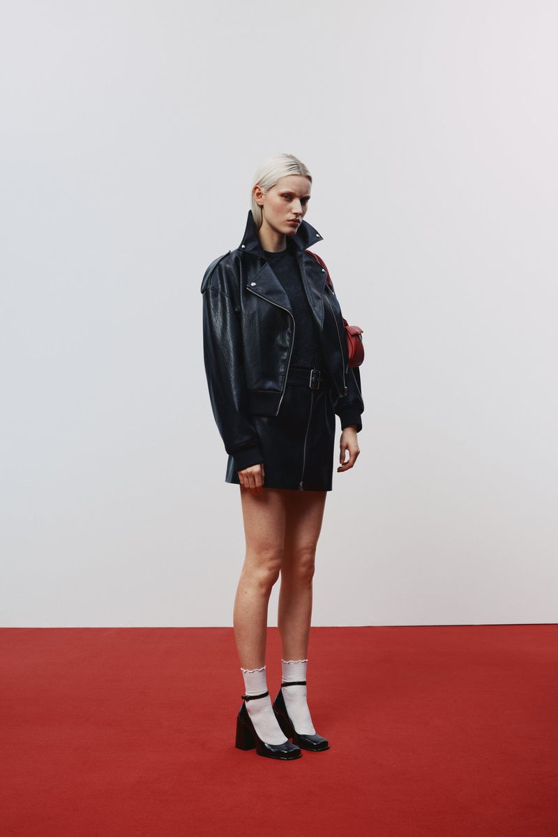 Full-shot of a blonde fashion model in a black leather jacket and skirt on a red carpet, with a white background in a studio setting.