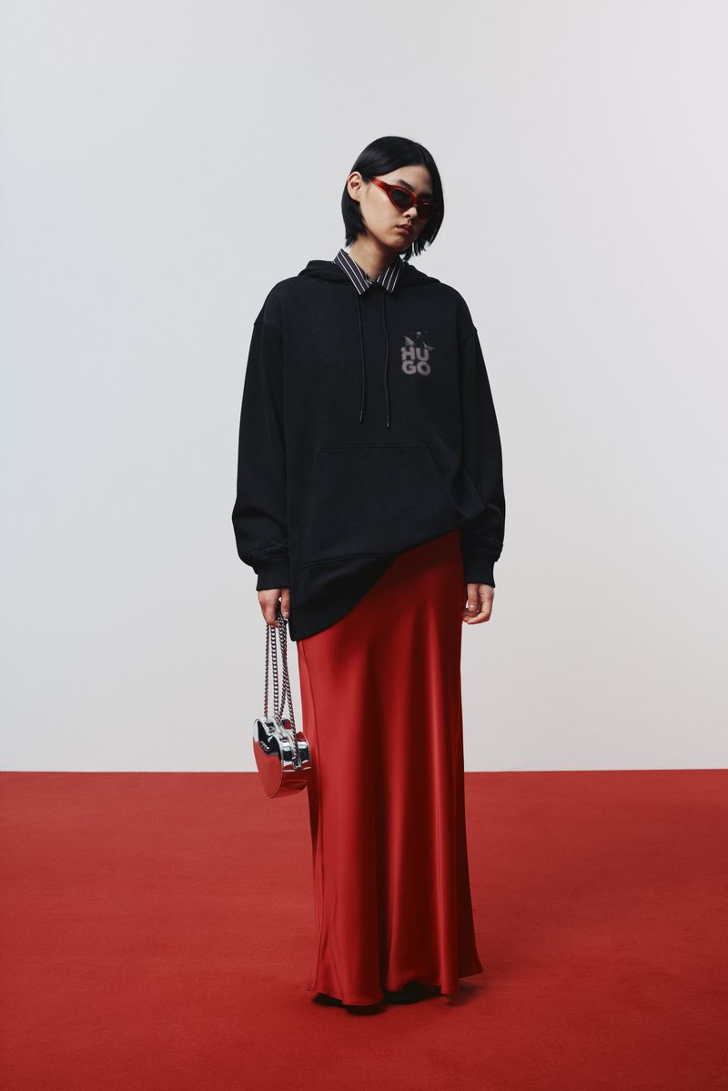 A person in a black hoodie and red skirt stands on a red surface in front of a white background holding a metallic purse.