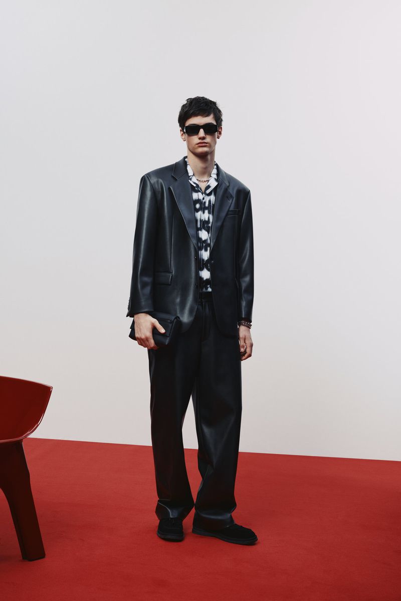 A man in a black suit and sunglasses poses on a red surface against a white backdrop, holding a black wallet, looking towards the camera.