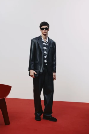 A man in a black suit and sunglasses poses on a red surface against a white backdrop, holding a black wallet, looking towards the camera.