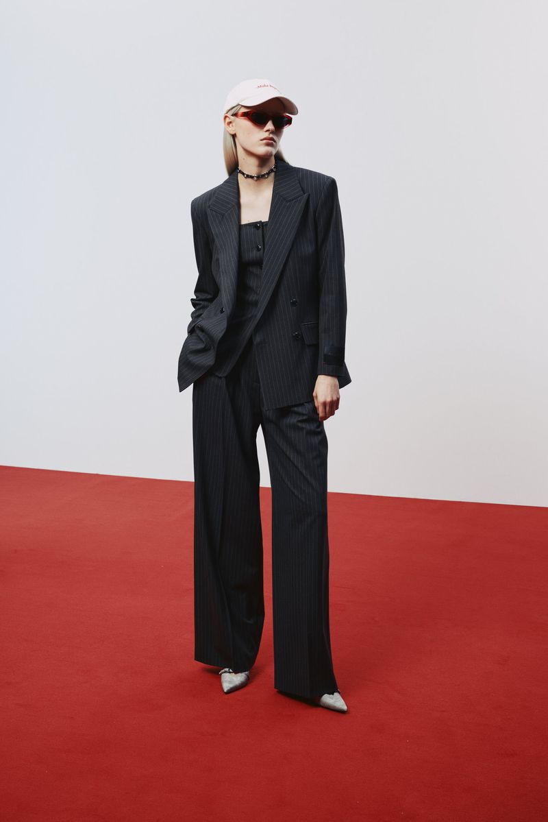 Woman in a pinstripe suit, pink cap, and sunglasses stands confidently on a red carpet against a white backdrop in a fashion editorial shot.