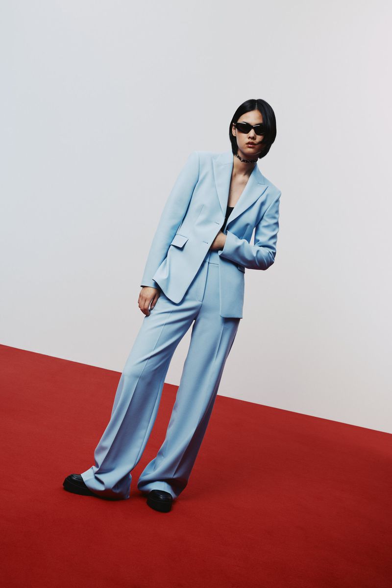 A woman in a light blue pantsuit with sunglasses poses on a red and white surface, creating a striking contrast in a studio setting.