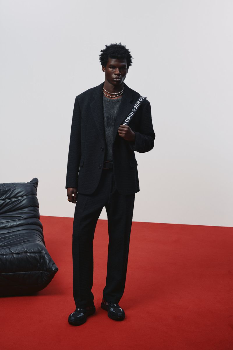 Man in a black suit stands on red floor against a white backdrop, featuring a black quilted chair and a branded crossbody bag.