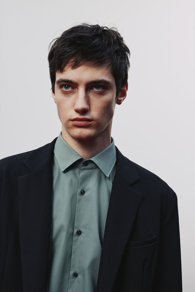 Close-up portrait of a young adult man in a black blazer and light green shirt against a white background. Studio lighting, fashion.