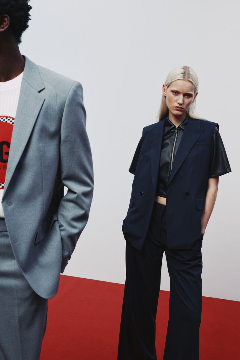 Two models showcasing contemporary fashion in a studio setting, one in a grey suit and the other in a short-sleeved navy blazer and pants.