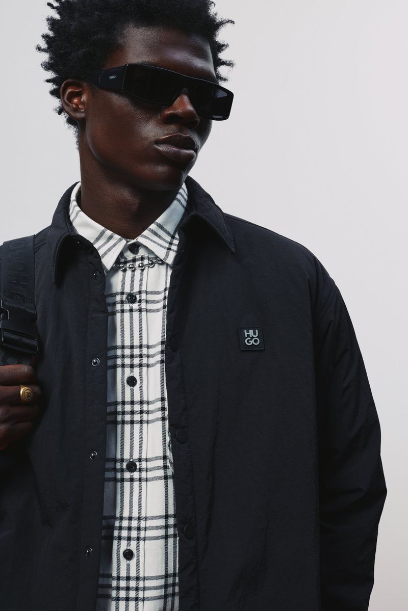 Black male model wearing dark sunglasses, an open black jacket, and a white plaid shirt against an off-white background.
