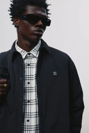 Black male model wearing dark sunglasses, an open black jacket, and a white plaid shirt against an off-white background.