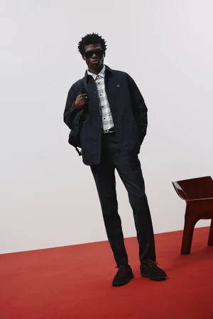 Model wearing dark jacket, plaid shirt, and sunglasses standing on a red floor against a white wall.