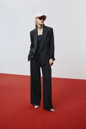 Model wearing an oversized dark pinstripe suit, pink baseball cap, and sunglasses on a red floor for HUGO.