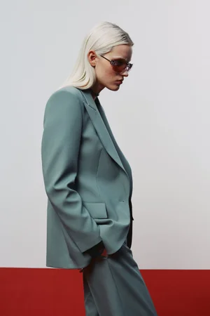 Model wearing a sage green suit jacket and tinted sunglasses against a white background with red floor