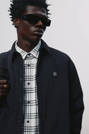 Black male model wearing dark sunglasses, an open black jacket, and a white plaid shirt against an off-white background.