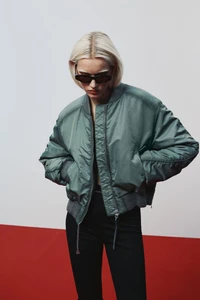 Model with blonde hair and dark sunglasses wearing an oversized sage green bomber jacket and black trousers on a red and white set.