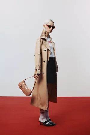 Model wearing a beige trench coat, sunglasses, and carrying a shoulder bag against a white and red studio background