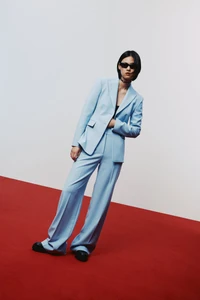 Model wearing a light blue tailored suit and black sunglasses posing on a red floor against a white studio background.
