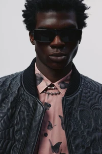 Black male model wearing dark rectangular sunglasses, an embroidered black leather jacket, and a pink butterfly shirt in studio.
