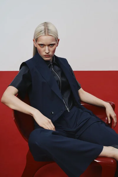 Model with blonde hair seated in a red chair wearing a navy blue vest and trousers against a dual toned background