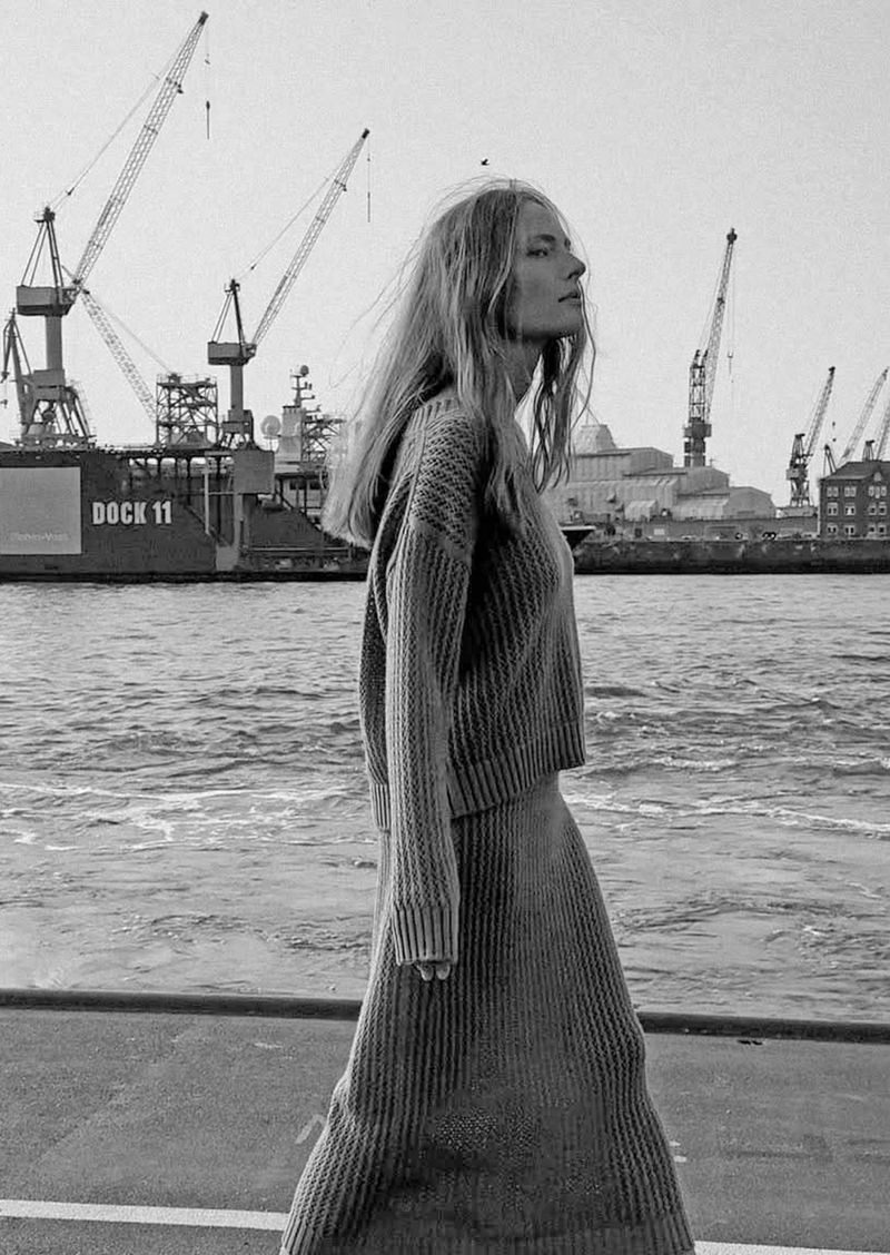 Monochrome fashion shot of a woman in a knit sweater and skirt walking near a harbor with cranes and a ship in the background.